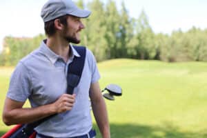 portrait of male golfer carrying golf bag while walking by green grass of golf club.